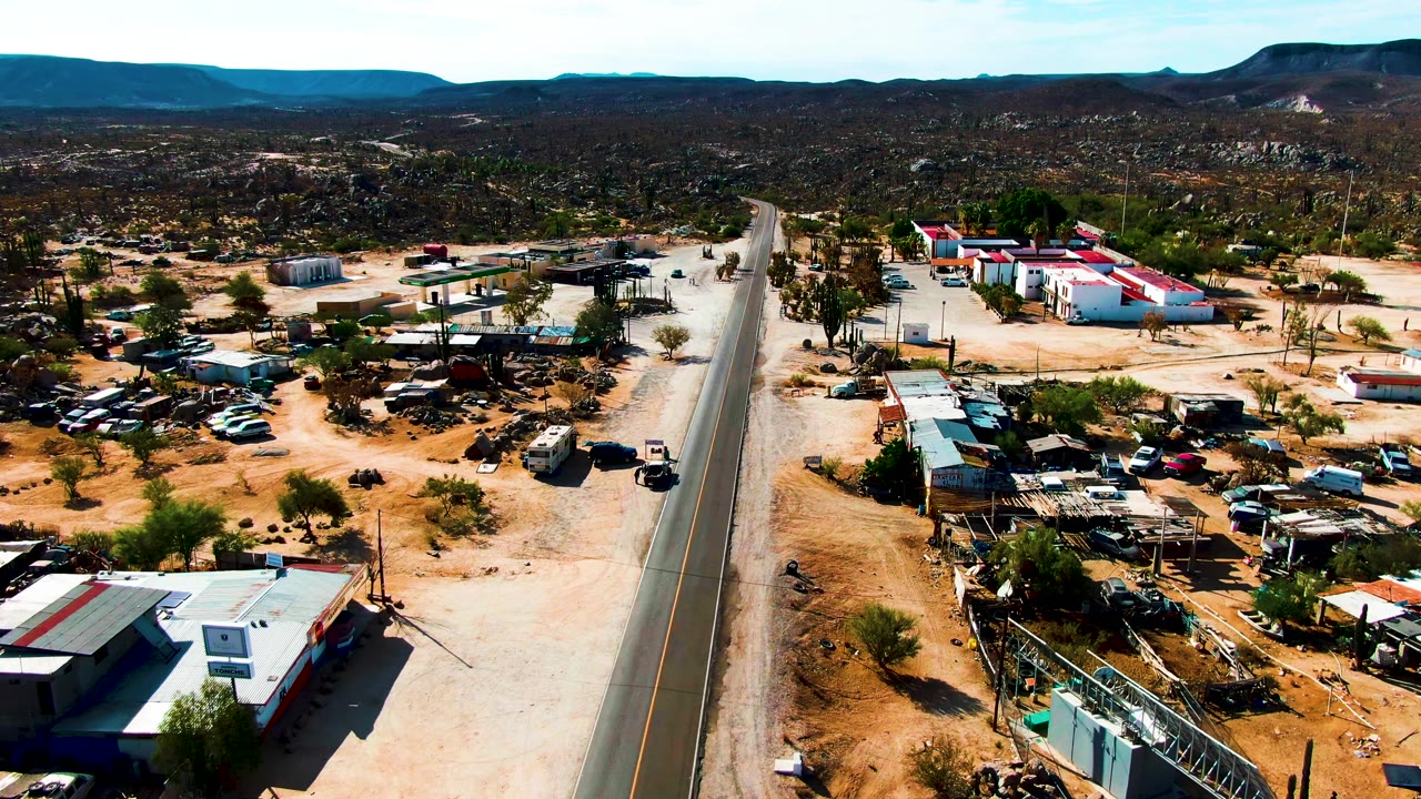 High aerial frame over Catavina BC Mexico showing a paved desert road, sandy lots, small buildings, parked vehicles, distant mountains, and no visible people.