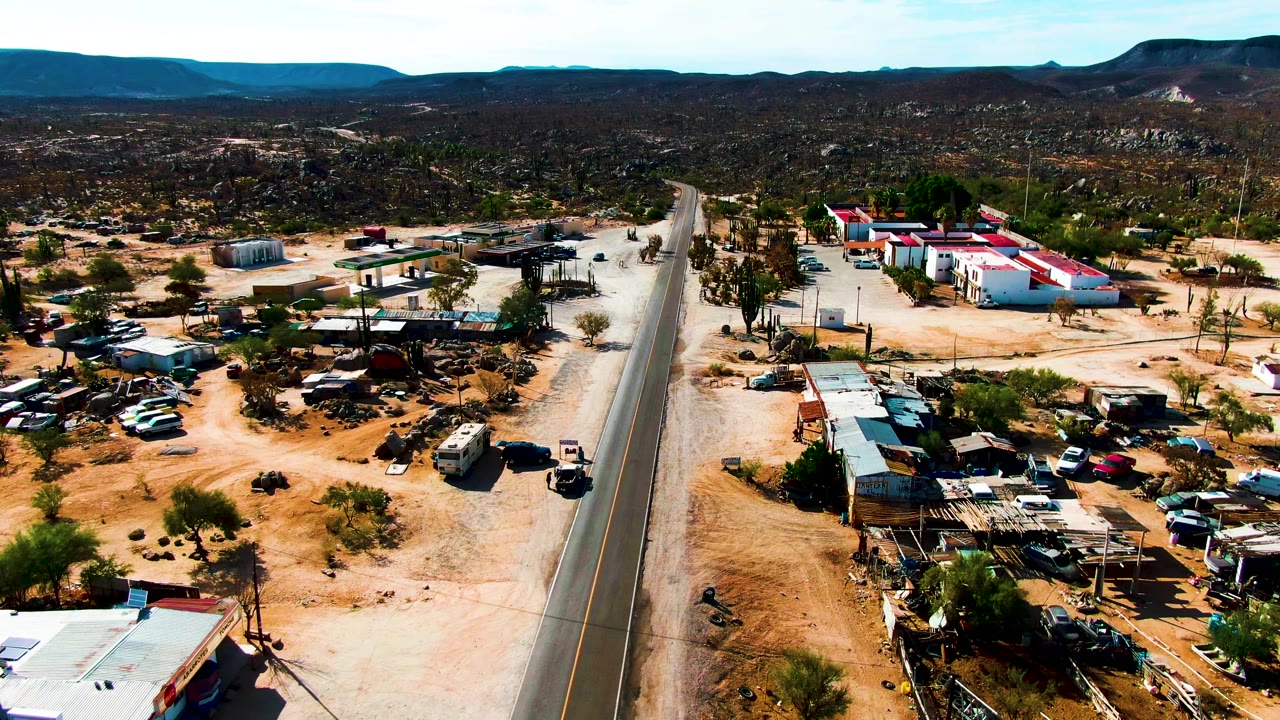 Aerial view in Catavina BC Mexico with the road centered near the frame, low roadside buildings, parked cars and trucks, desert vegetation, distant hills, and no visible people.