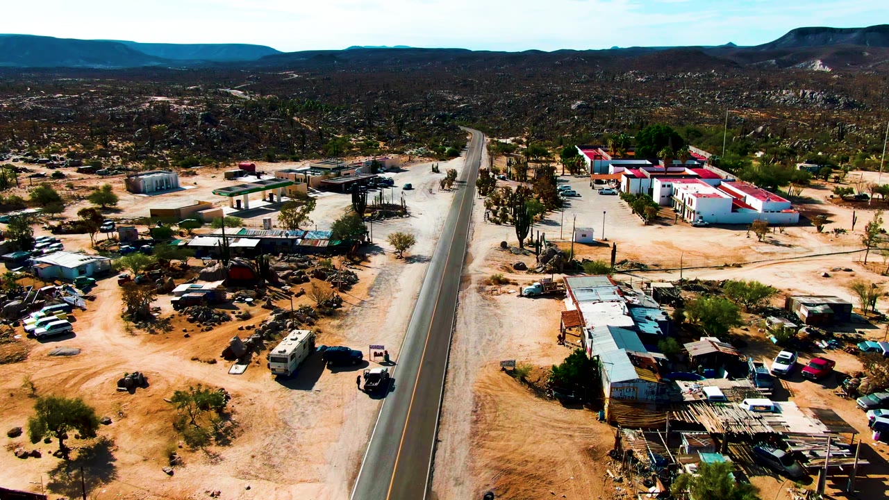 Catavina BC Mexico aerial frame showing white buildings with red roofs to the right of the road, parked vehicles in sandy lots, cacti, low mountains, and no visible people.