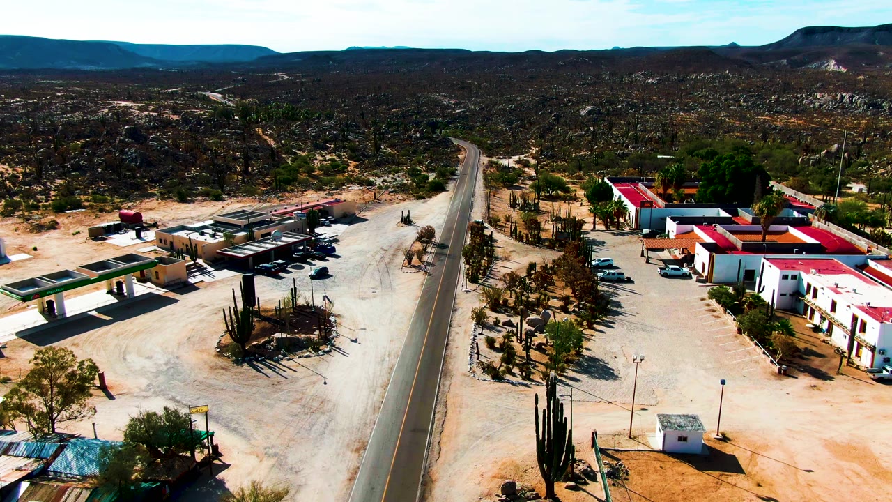 Aerial frame over Catavina BC Mexico showing a paved road, sandy lots, low buildings with red roofs, tall cacti, distant mountains, and no visible people.