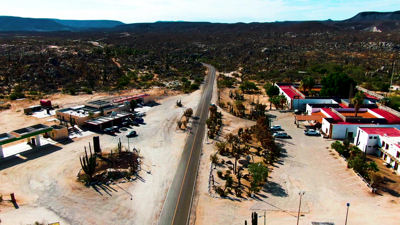 Aerial view in Catavina BC Mexico with the road running up the left side, red-roofed buildings on the right, cactus landscaping, parked vehicles, and no visible people.
