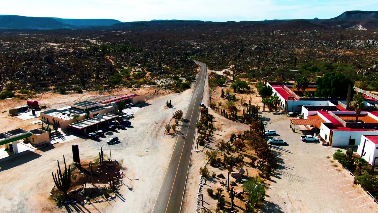Drone frame over Catavina BC Mexico showing a broad sandy roadside area, a central paved road, white and tan buildings, parked cars, cacti, dark hills, and no visible people.