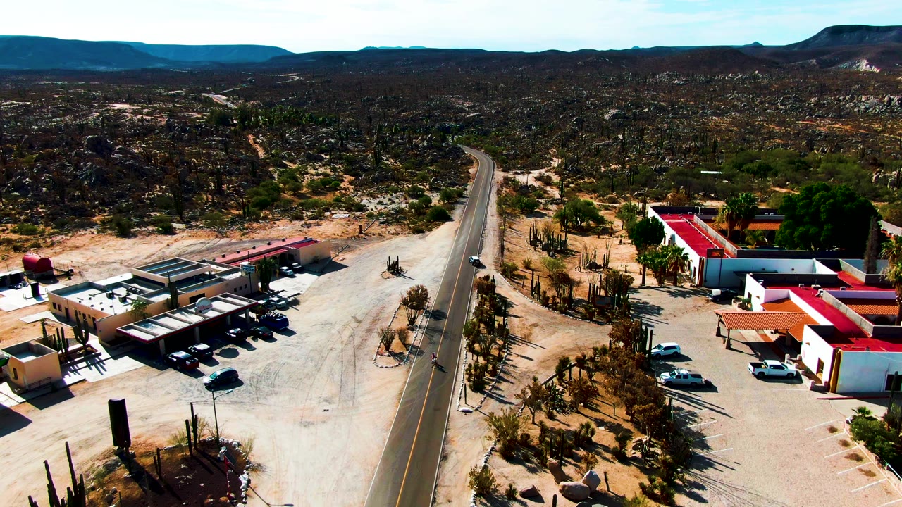 Aerial view in Catavina BC Mexico where a small vehicle is visible on the paved road, with red-roofed buildings, desert cacti, gravel lots, mountains, and no visible people.
