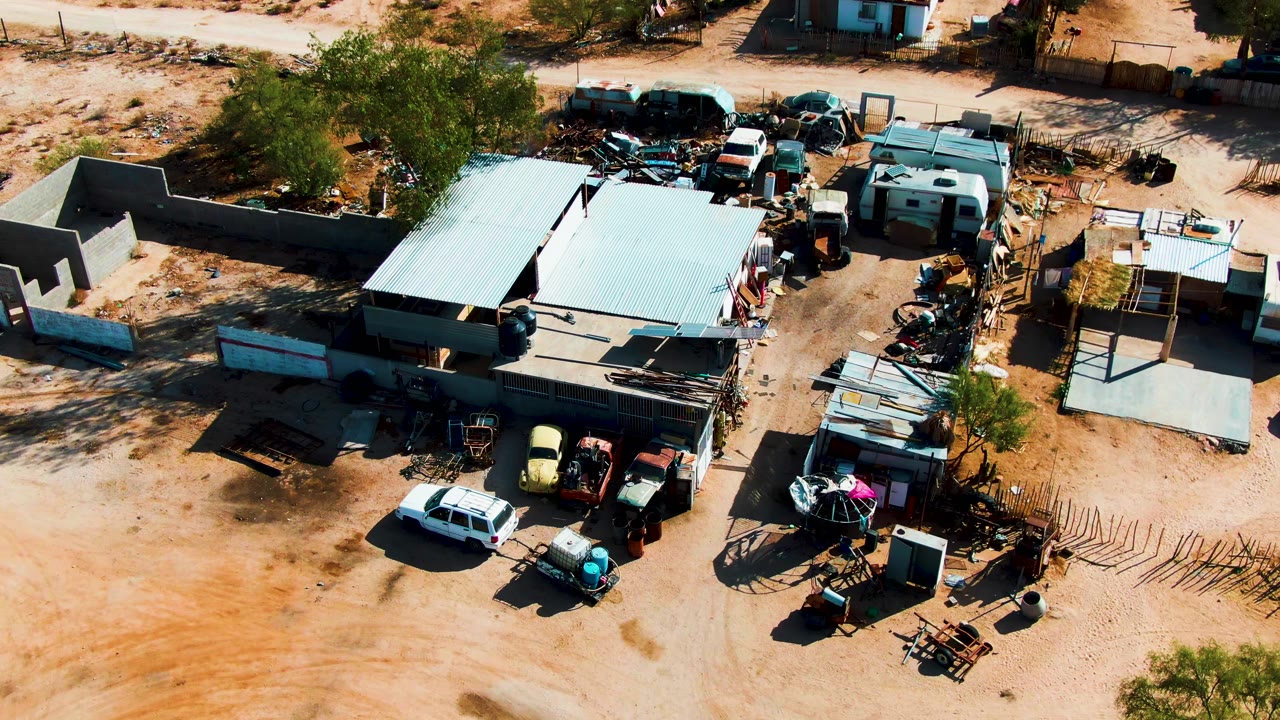 Opening overhead frame of a Catavina, Mexico junkyard with corrugated roofs, old cars, trailers, scrap piles, fencing, sparse trees, and no visible people.