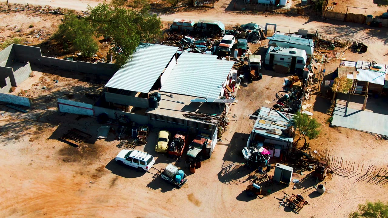 Early aerial frame over the Catavina, Mexico yard, showing a white car near the lower edge, trailers at the upper right, stacked metal, shade from trees, and no visible people.