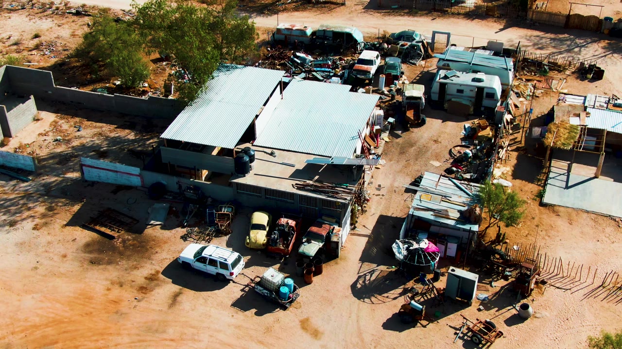 Aerial frame in Catavina, Mexico with the main metal-roofed structure across the center, trailers and scrap at the top, a fenced yard to the right, and no visible people.