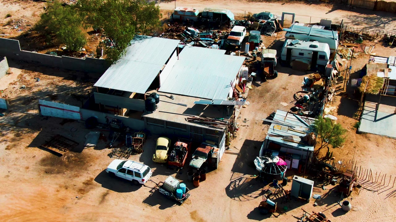 Closer overhead view of metal roofs, old vehicles, scrap piles, and dusty ground.