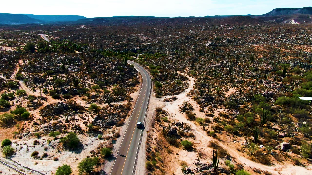 A dark car travels along a paved road through rocky cactus desert near Catavina, Mexico, with distant mountains and no visible people.
