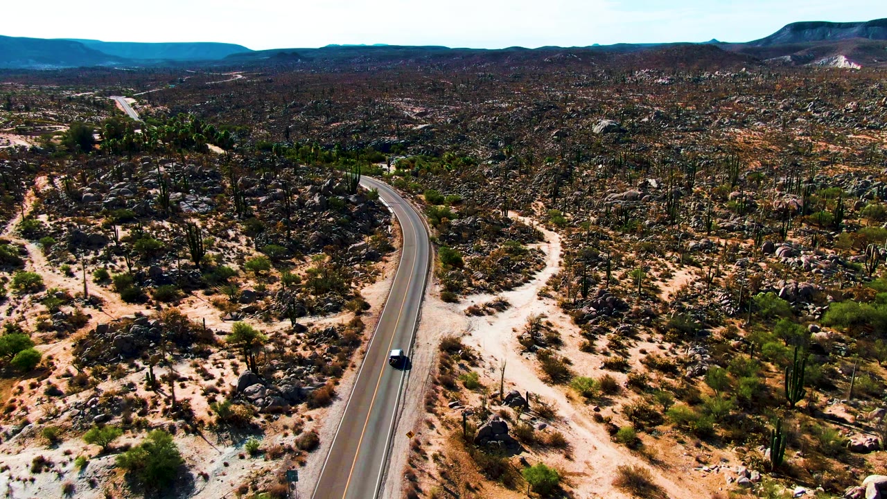 The car continues along the curving road through Catavina desert terrain, with cactus clusters, pale sand tracks, dark rocks, and no visible people.