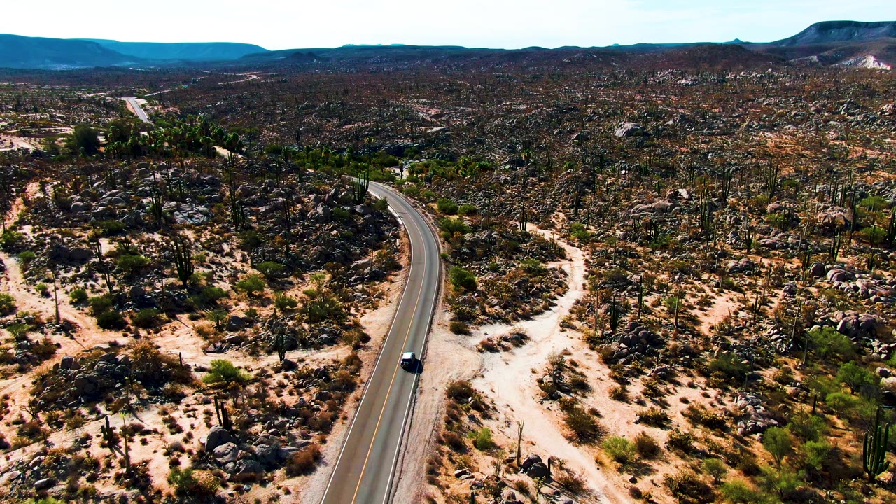 A high aerial frame shows the car on the road beside scattered cacti and boulder fields in Catavina, Mexico, with a low mountain horizon and no visible people.