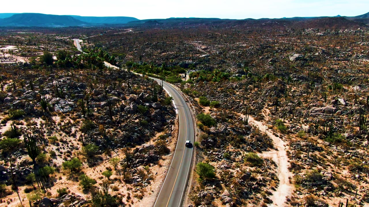 Aerial view of a winding desert road with a small car and rocky cactus terrain.