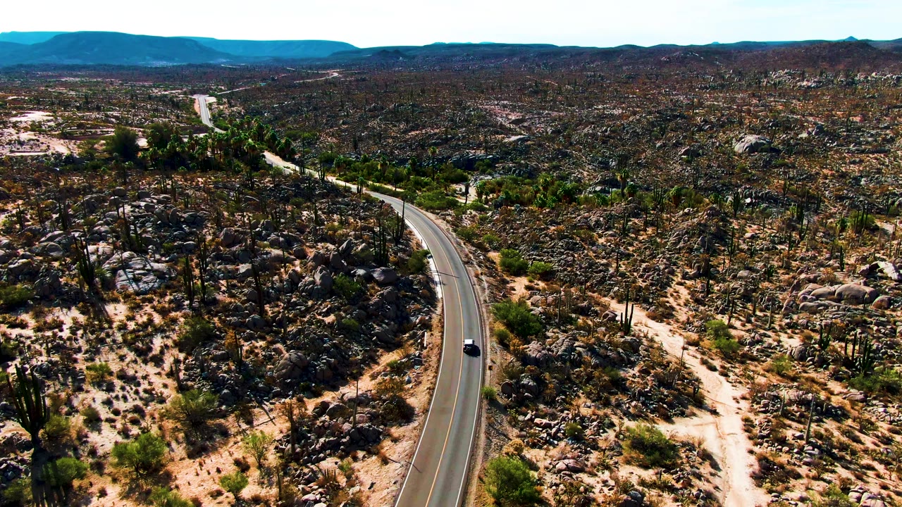 At 0:01, the car appears farther along the lower bend of the road as the highway cuts past boulders, cacti, pale sand tracks, and no visible people in Catavina BC Mexico.