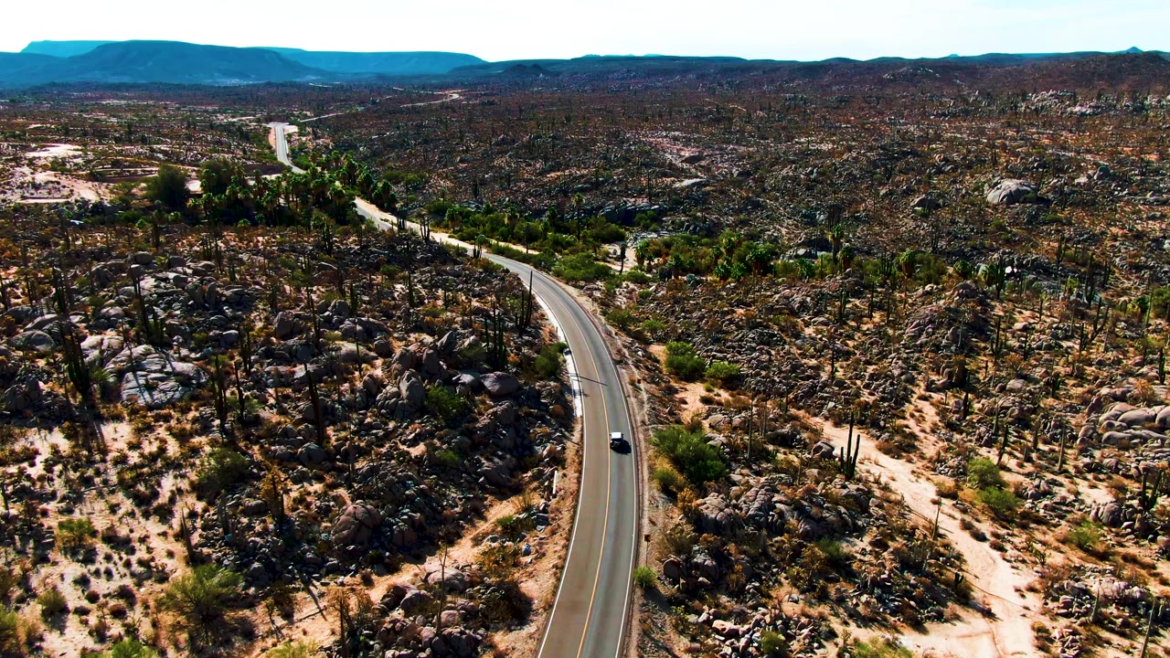 At 0:02, a wide aerial frame places the car on the right-side curve of the Catavina BC Mexico road, surrounded by dark rocks, scattered cacti, dry washes, and no visible people.
