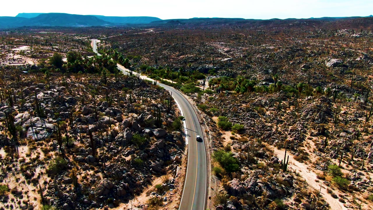 At 0:03, the paved road bends up through the frame with the small car near the lower section, while rocky cactus terrain and a low mountain horizon fill Catavina BC Mexico with no visible people.