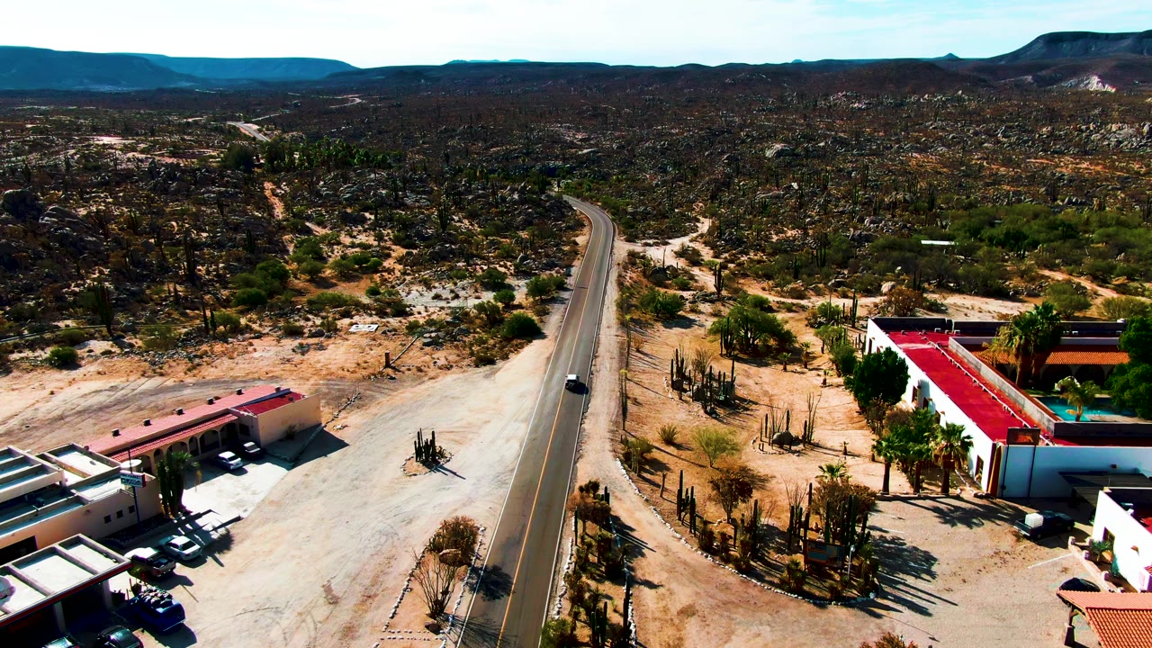Aerial frame from Catavina BC, Mexico showing a paved desert road, red-roofed roadside buildings, cactus clusters, sandy ground, distant mountains, and no visible people.