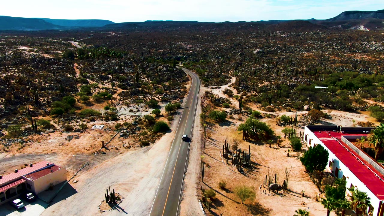 Catavina BC, Mexico aerial frame showing the road running into the desert, a small dark car, a white building with a red roof at lower right, cactus stands, sandy washes, and no visible people.