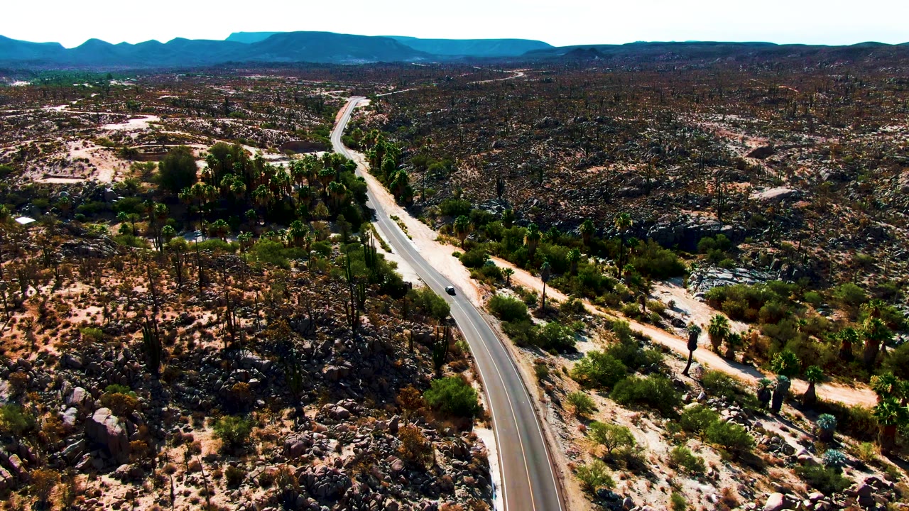 Aerial frame over Catavina BC, Mexico, showing a paved highway entering from the lower center through rocky cactus desert toward distant mountains, with no visible people.