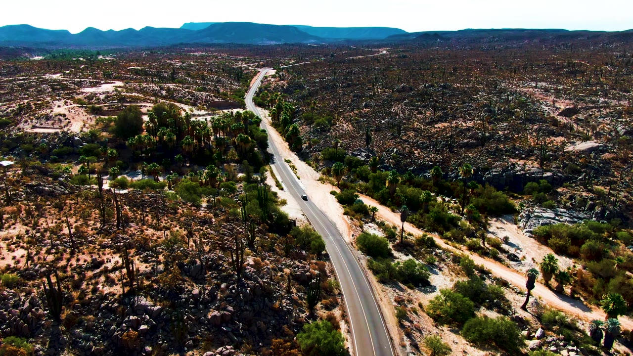 Aerial frame over Catavina BC, Mexico, showing a small vehicle on the paved road, dark rock outcrops, scattered cacti, and a long mountain horizon with no visible people.