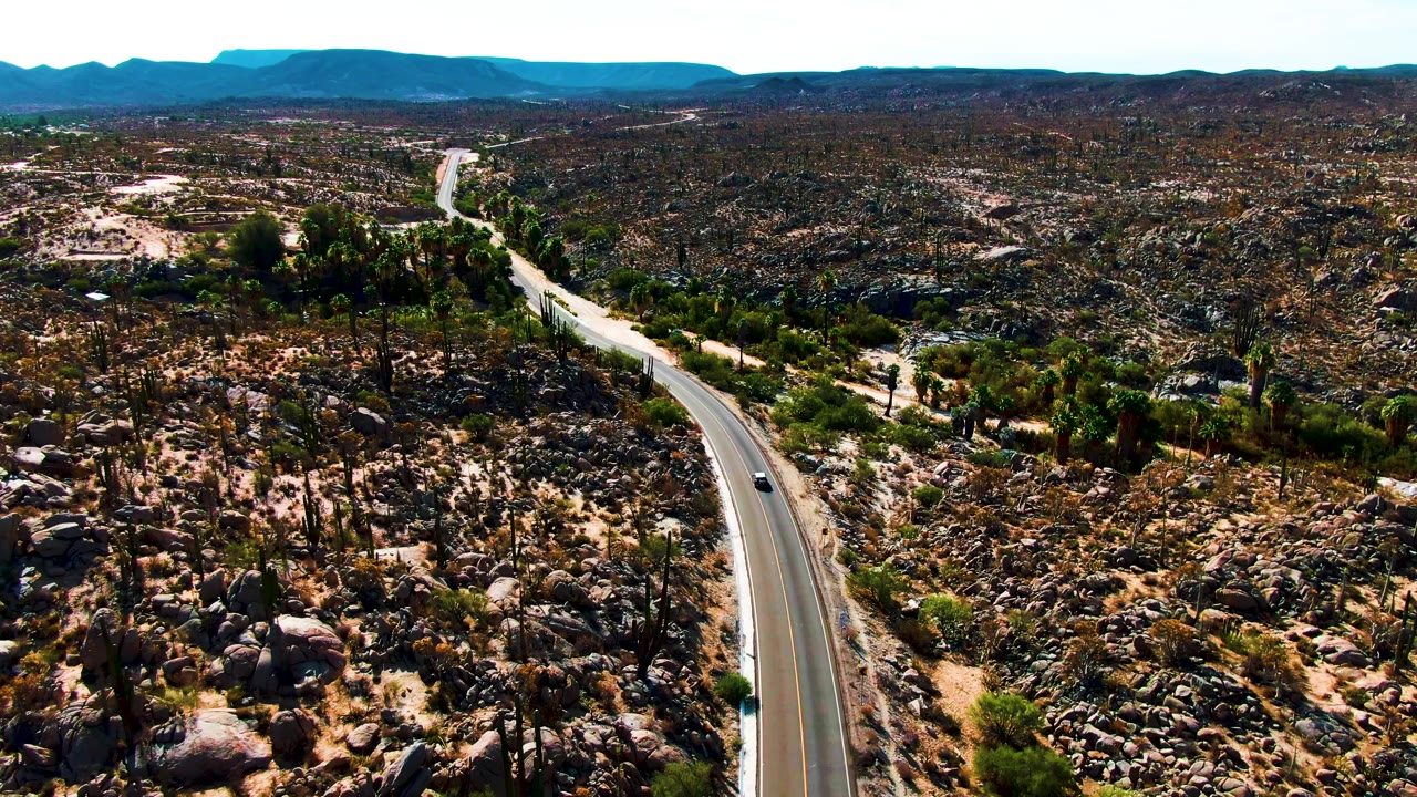 Aerial view in Catavina BC Mexico with a small car on the lower curve of a paved desert road, rocky cactus terrain on both sides, distant mountains, and no visible people.