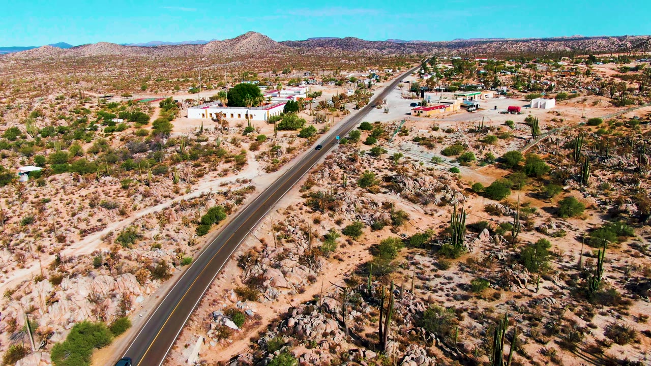 Aerial frame from Catavina BC, Mexico showing a dark car on a two-lane desert road beside rocky ground, tall cacti, low buildings, distant hills, blue sky, and no visible people.