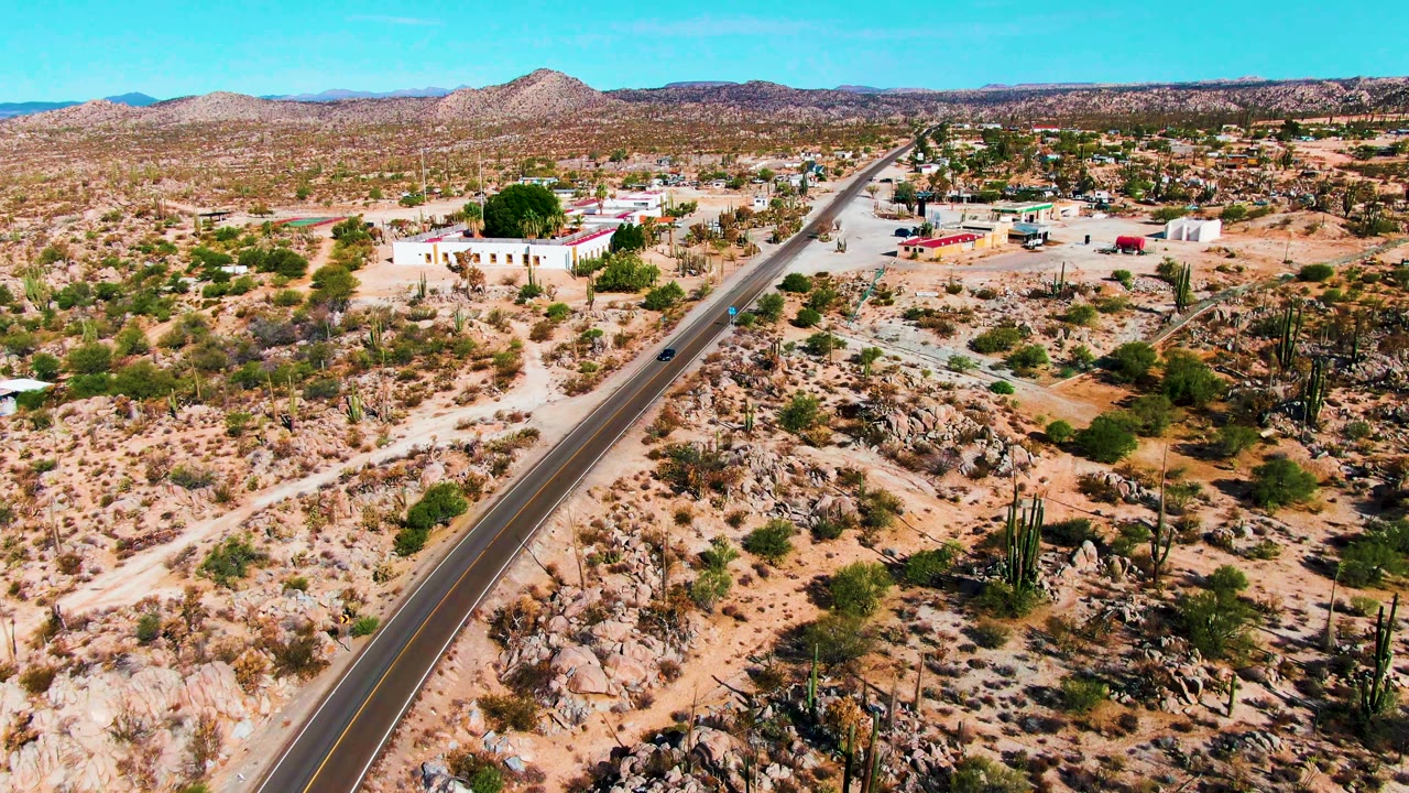 Aerial view in Catavina BC, Mexico with the road running diagonally past cactus-covered rocky ground, pale buildings, scattered vehicles or structures in the distance, and no visible people.