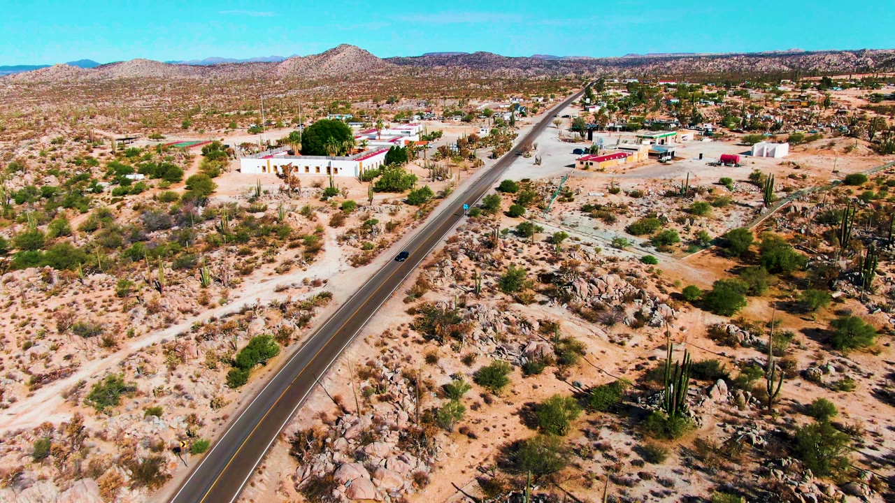 Drone-height frame from Catavina BC, Mexico showing the paved road cutting through rocky desert, a car on the roadway, clusters of cacti, scattered buildings, low mountains, and no visible people.