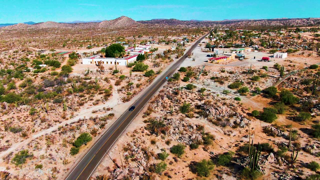 Wide aerial view of a car on a desert road near cacti, rocks, and scattered buildings.