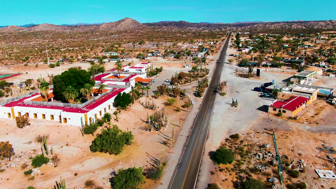 Aerial view in Catavina BC Mexico showing a paved road running past cream buildings with red roofs, sandy lots, cacti, distant hills, blue sky, and no visible people.
