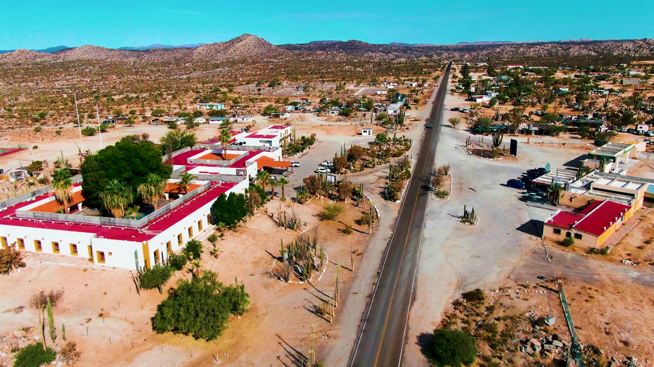 Drone view in Catavina BC Mexico with the red-roofed buildings shifted closer to center, a straight paved road at the right, sandy ground, desert plants, parked vehicles, and no visible people.