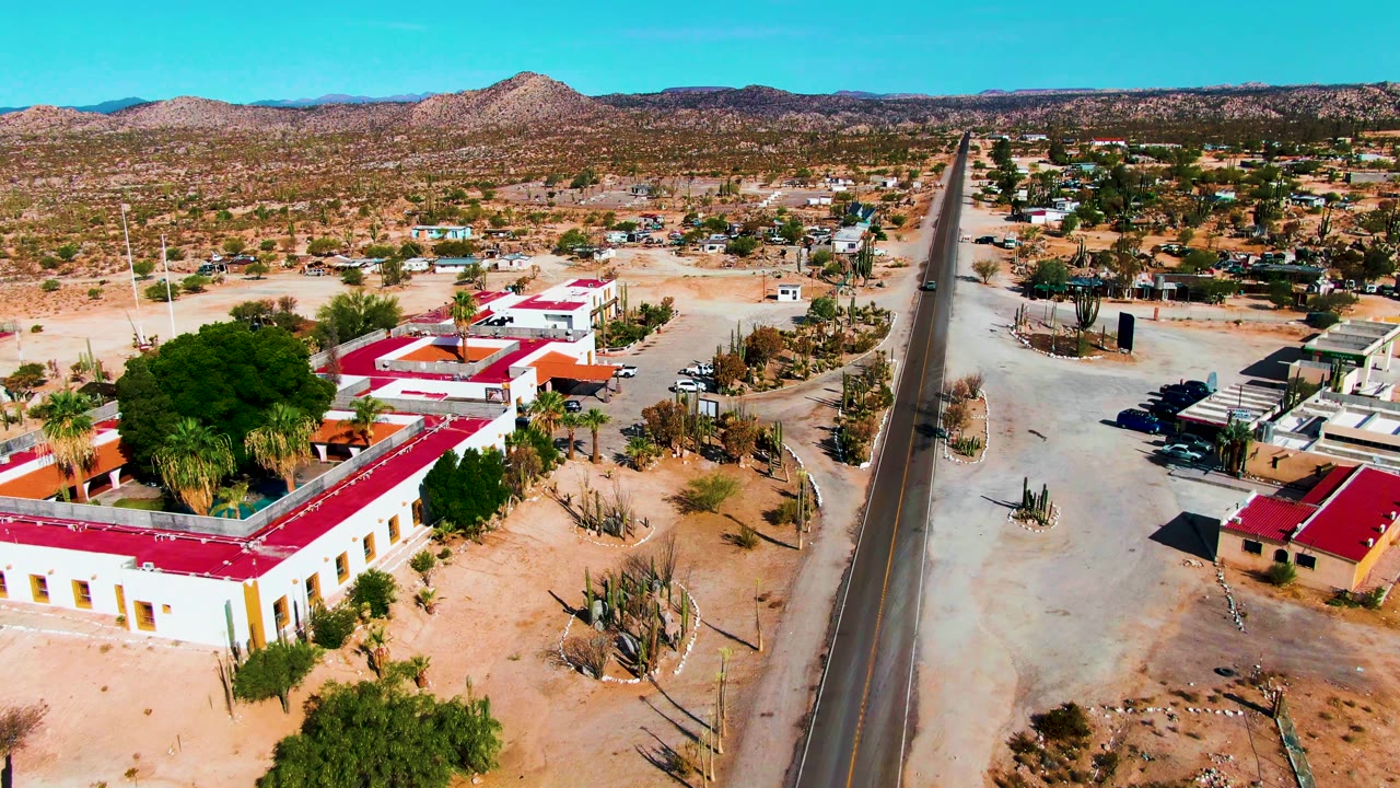 Aerial pass in Catavina BC Mexico showing cream buildings with red roofs, a roadside parking area, cacti, scattered trees, open sandy terrain, low mountains, and no visible people.