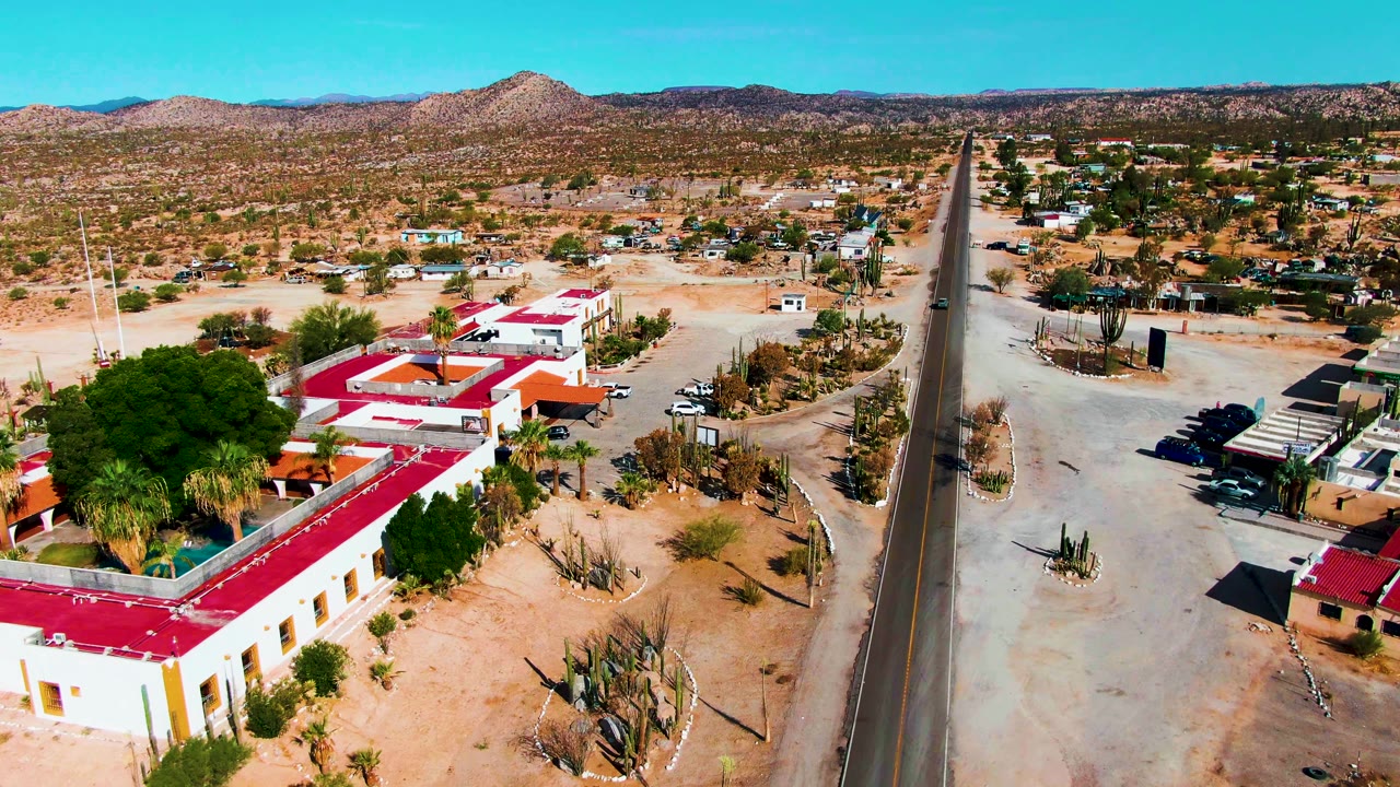 Wide aerial view of a desert road, buildings, cacti, and distant hills.