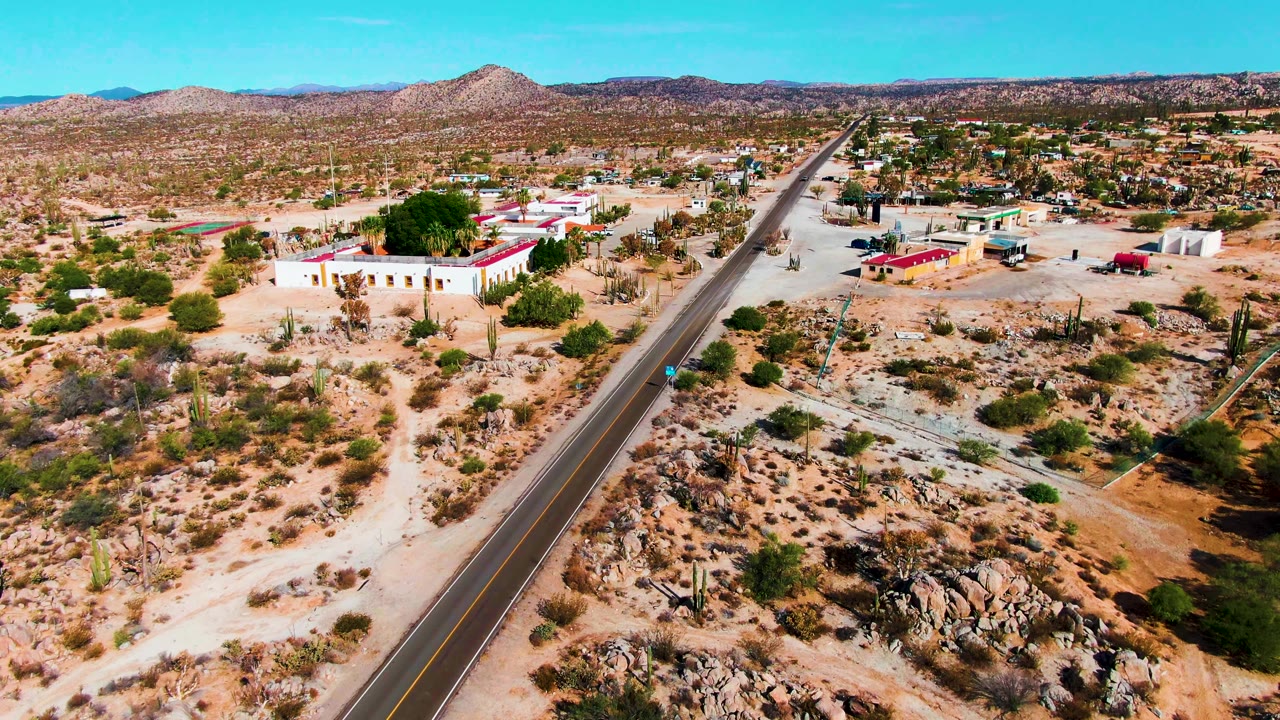 Early frame from Catavina BC Mexico showing a two-lane road through sandy desert, red-roofed buildings to the left, scattered cacti, distant hills, and no visible people.