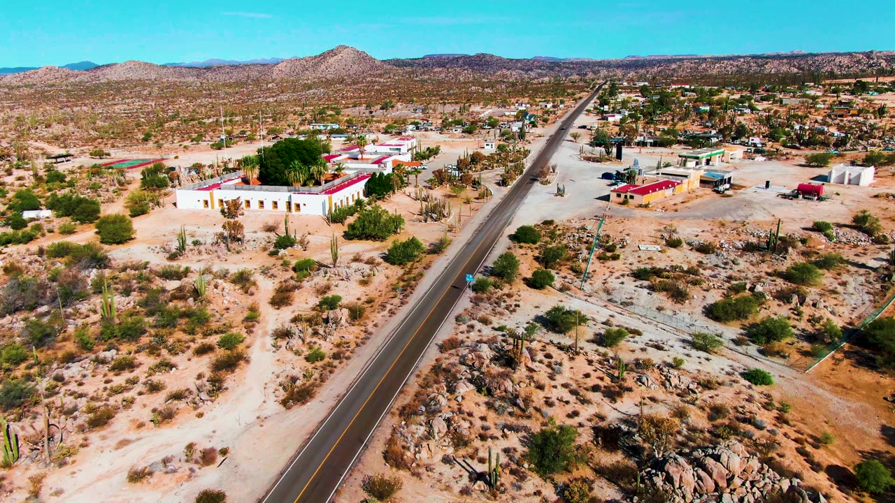 One second in, the Catavina BC Mexico road runs toward the horizon with white and red-roofed buildings, scrub, cacti, rocky patches, and no visible people.