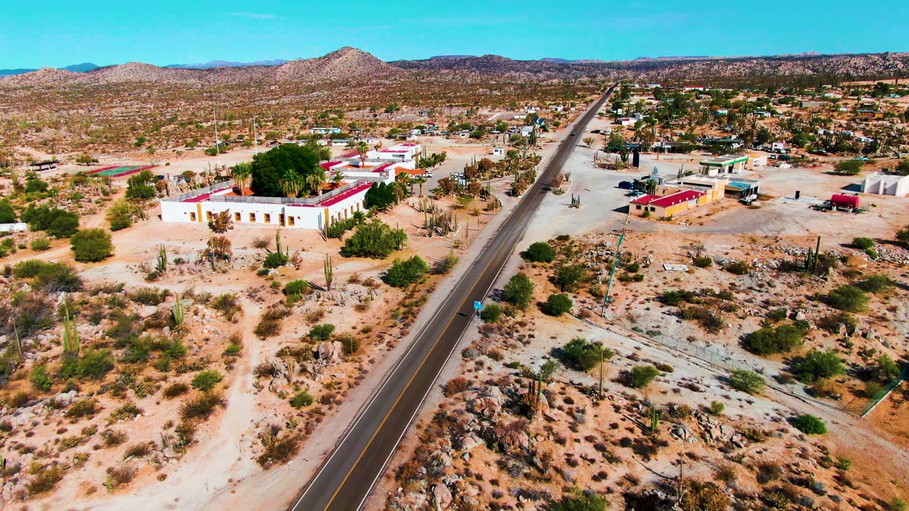 At 0:02, the aerial angle shows the road slightly right of center, desert buildings on both sides, cacti and scrub in the foreground, distant hills, and no visible people.