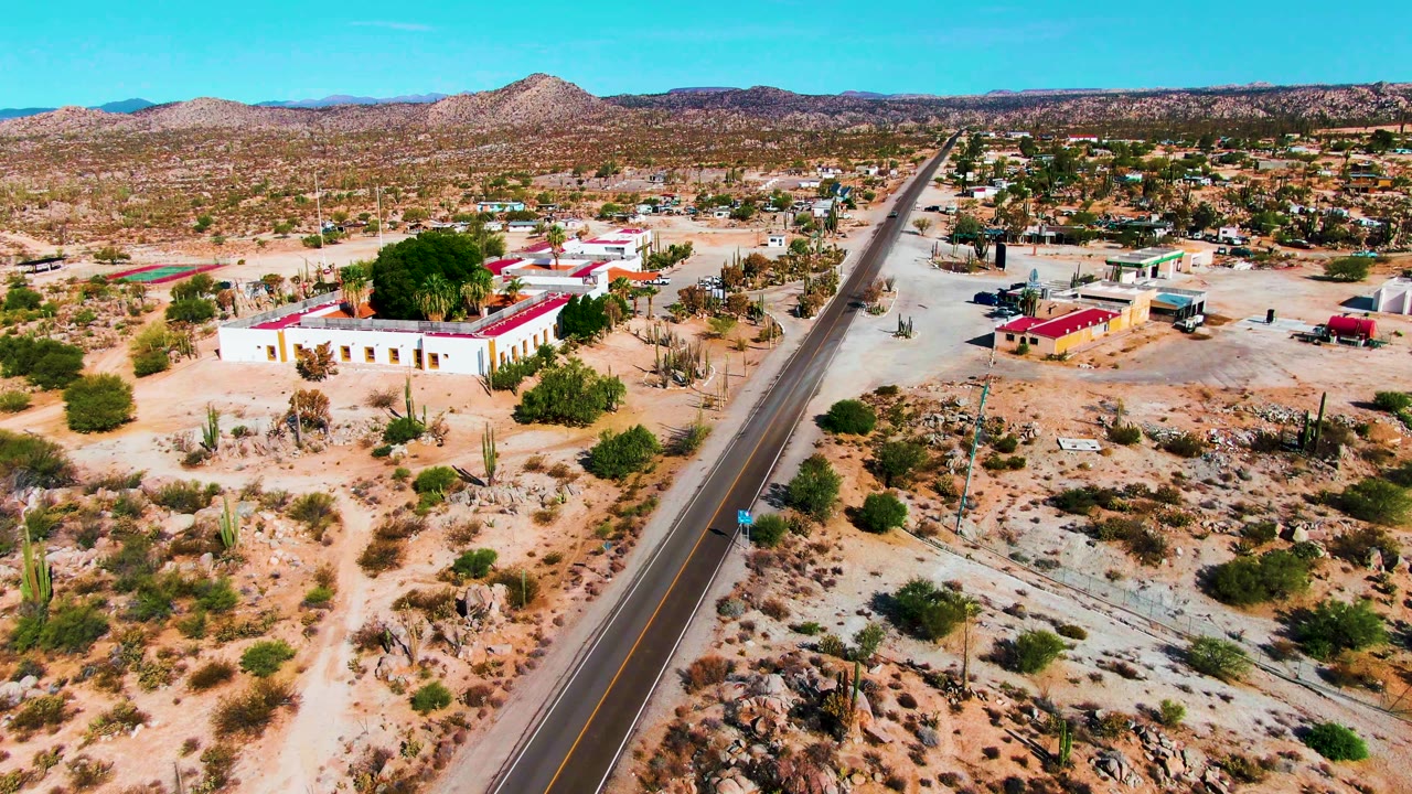 Aerial frame of a desert highway passing small buildings and cactus-covered ground.