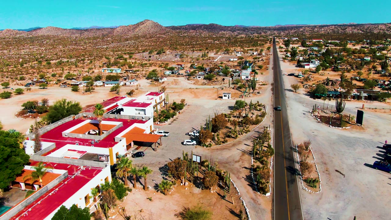 Aerial view in Catavina BC, Mexico showing red-roofed white buildings beside a straight desert road, sandy lots, cacti, distant hills, and no visible people.