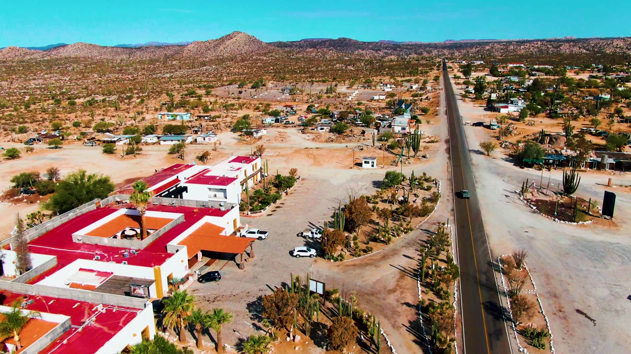 Drone view in Catavina BC, Mexico with white buildings and red roofs lower in frame, a straight paved road at center-right, scattered cacti, sandy terrain, distant mountains, and no visible people.