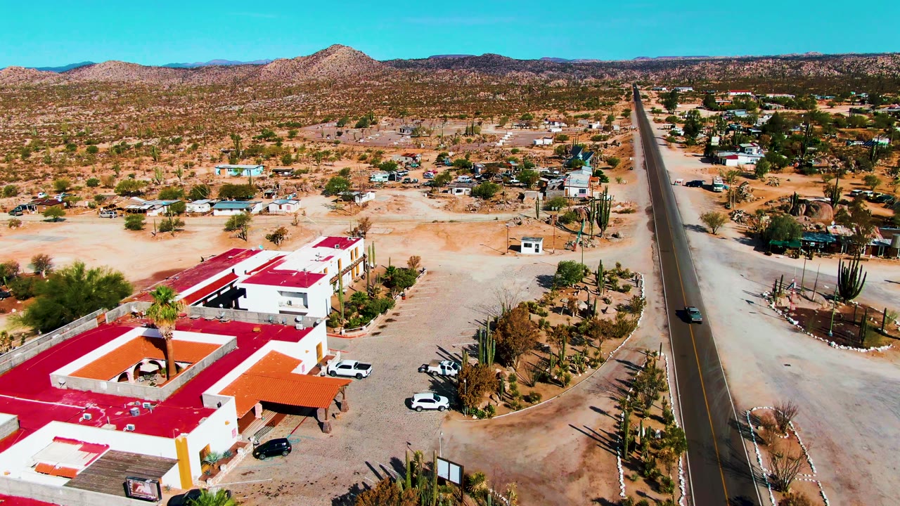 Aerial frame in Catavina BC, Mexico showing red-roofed roadside buildings, a paved road with a vehicle, sandy open ground, cactus clusters, low mountains, and no visible people.