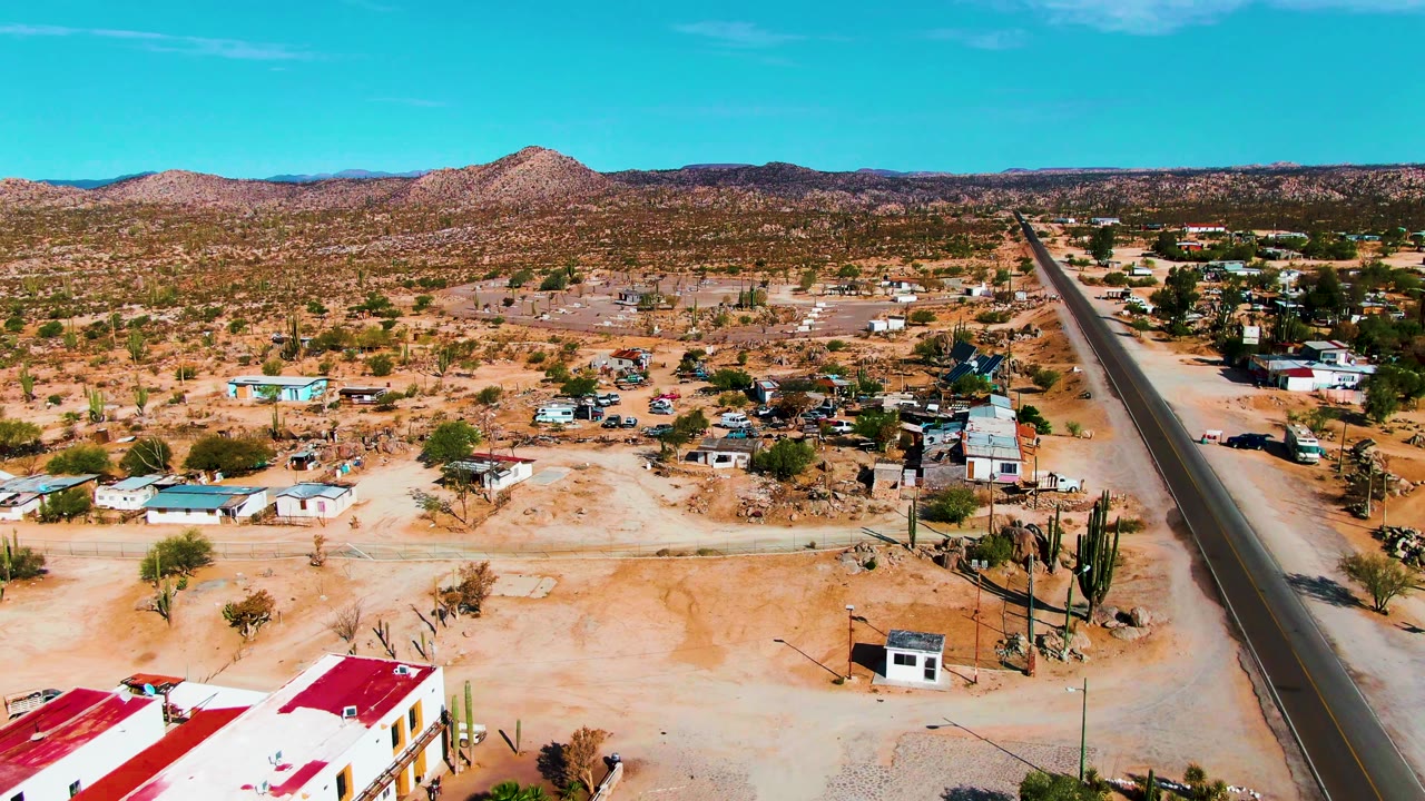 Aerial view in Catavina BC, Mexico showing a paved road on the right, low houses across dry orange ground, tall cacti near the road, distant rocky hills, blue sky, and no visible people.