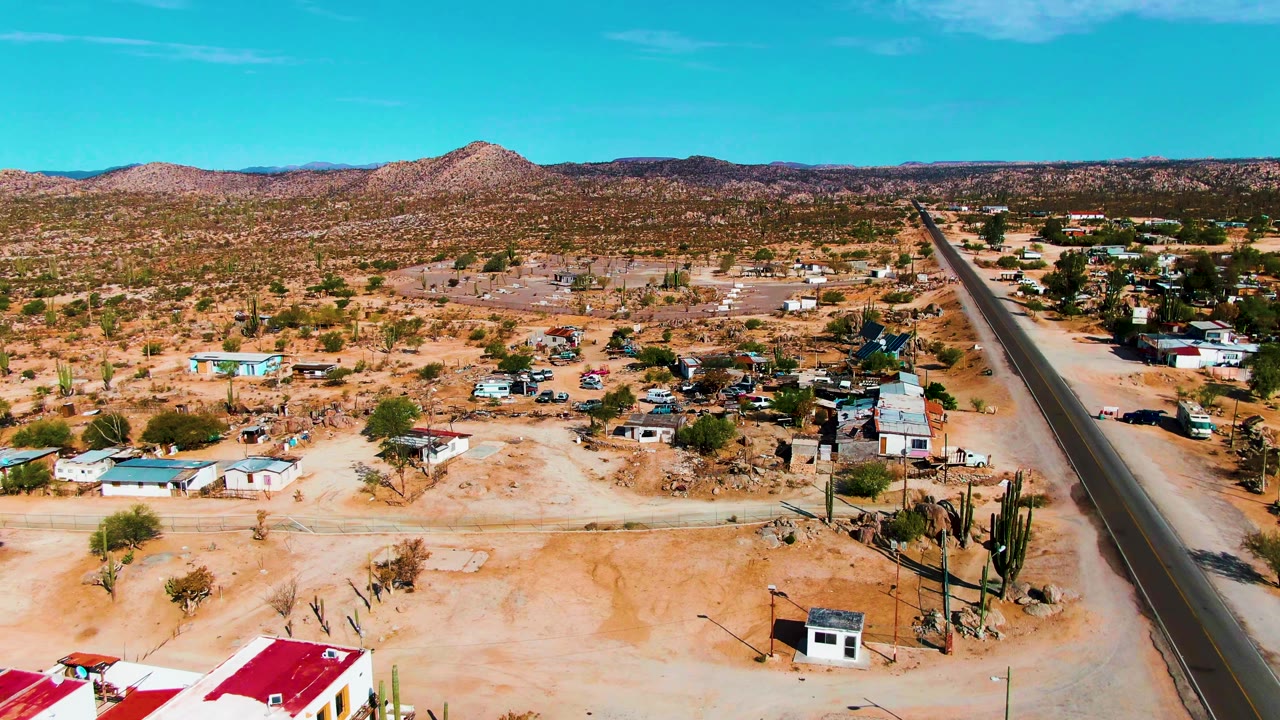 Desert settlement with a paved road, low buildings, cacti, and hills.