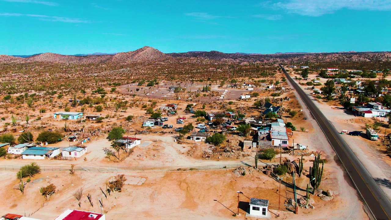 Aerial view of Catavina BC, Mexico with a straight paved road on the right, scattered desert homes and vehicles or small objects near the buildings, cacti in the lower right, low hills on the horizon, and no visible people.
