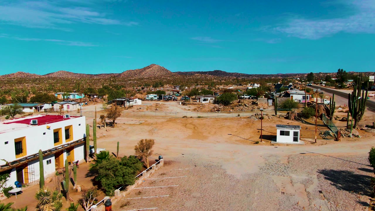 At 0:00, an elevated view in Catavina BC Mexico shows white low buildings at the lower left, tall cacti near the center, sandy ground, distant hills, blue sky, and no visible people.