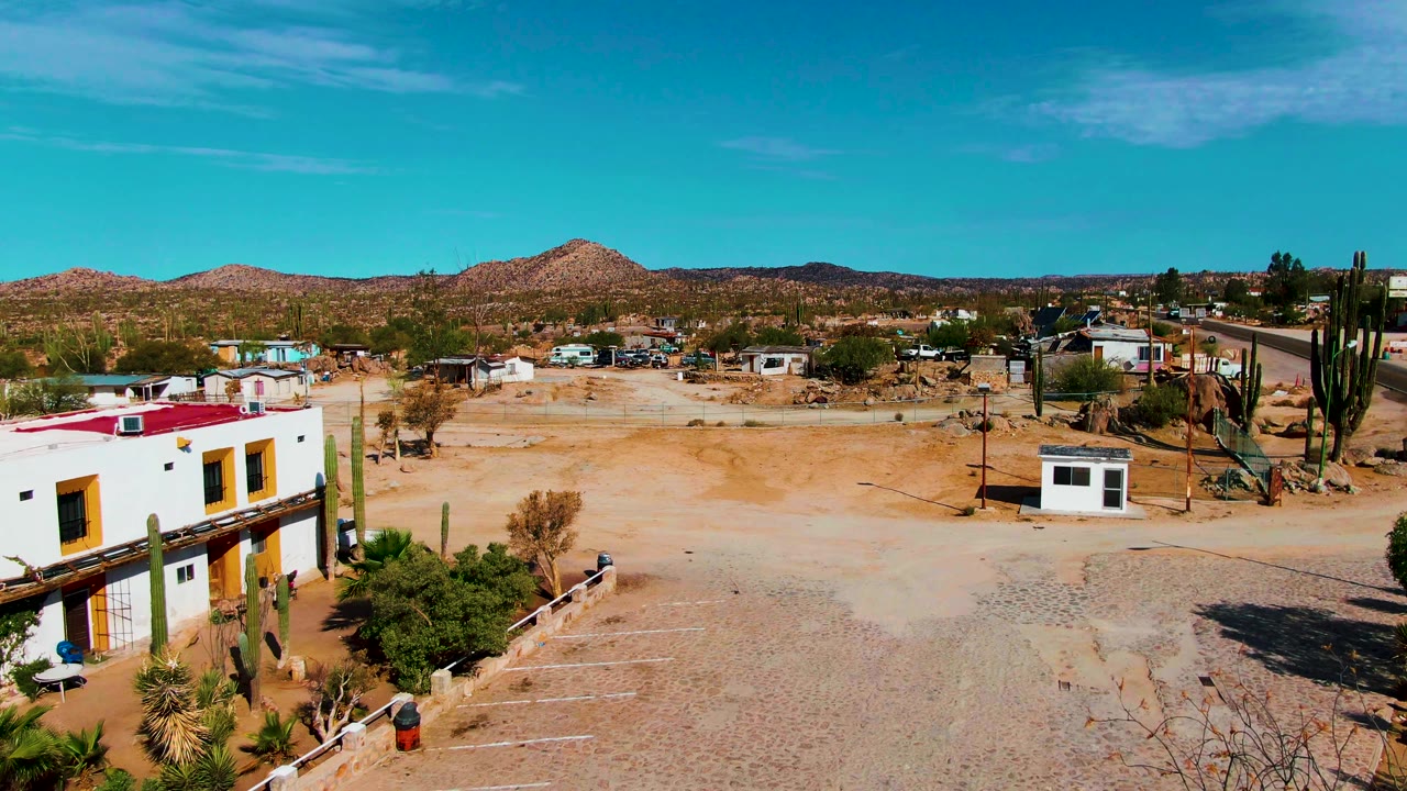 White building with yellow trim beside cacti and open sandy desert ground.