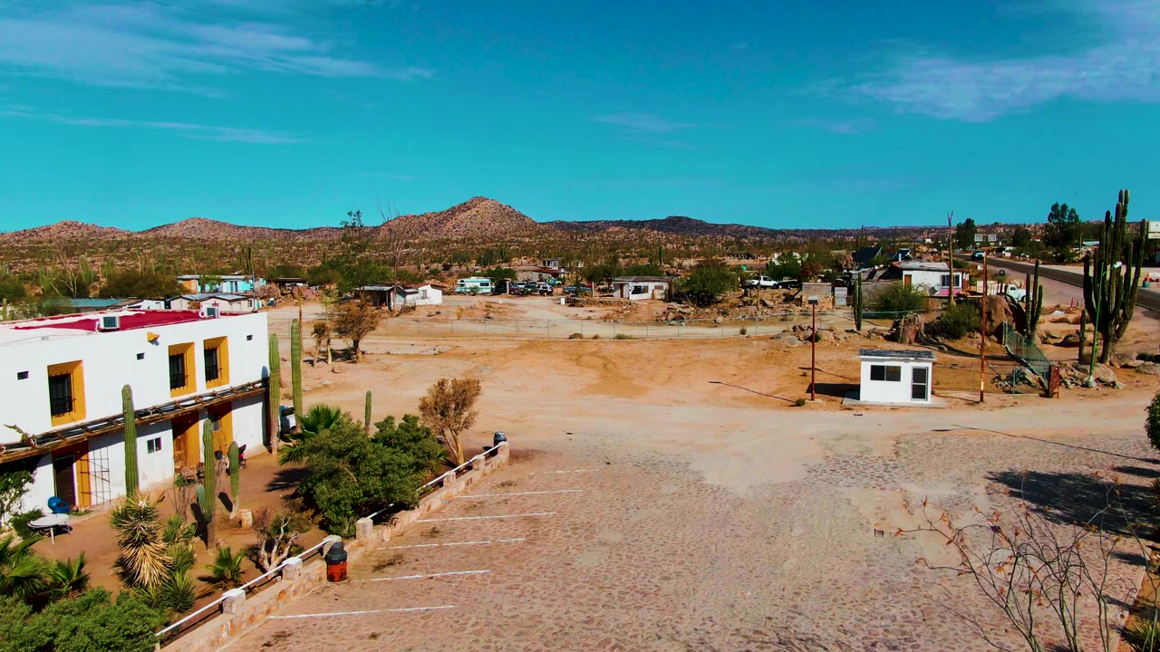 At 0:02, the frame looks across sandy ground in Catavina BC Mexico toward a small white structure, tall cacti, low buildings, distant hills, clear sky, and no visible people.