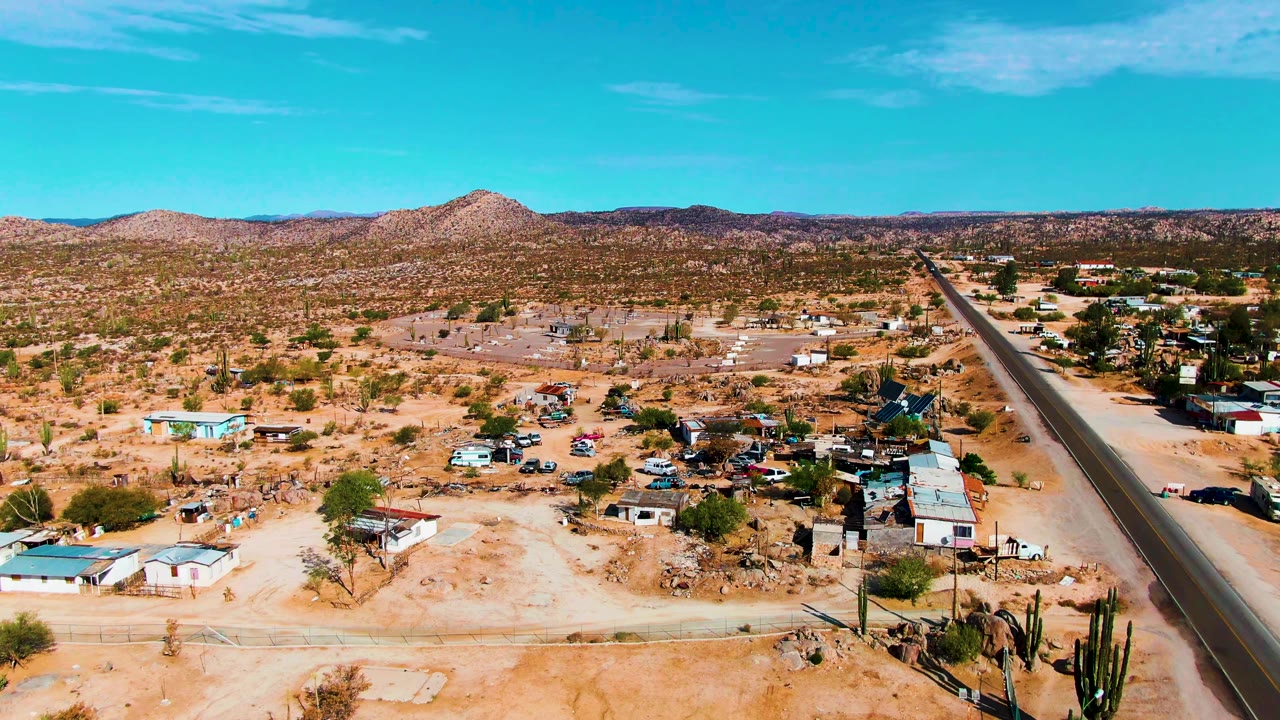 Aerial frame over Catavina, Mexico showing sandy lots, scattered small buildings, tall cacti near the lower edge, a paved road at right, distant hills, and no visible people.
