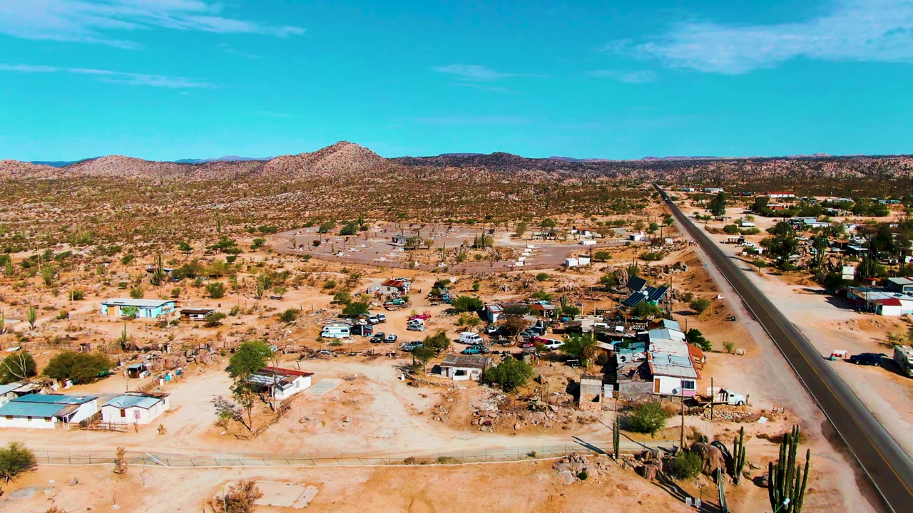Aerial frame in Catavina, Mexico with the paved road running from foreground to distance, scattered homes and sheds on sandy ground, cactus clusters, mountains on the horizon, and no visible people.