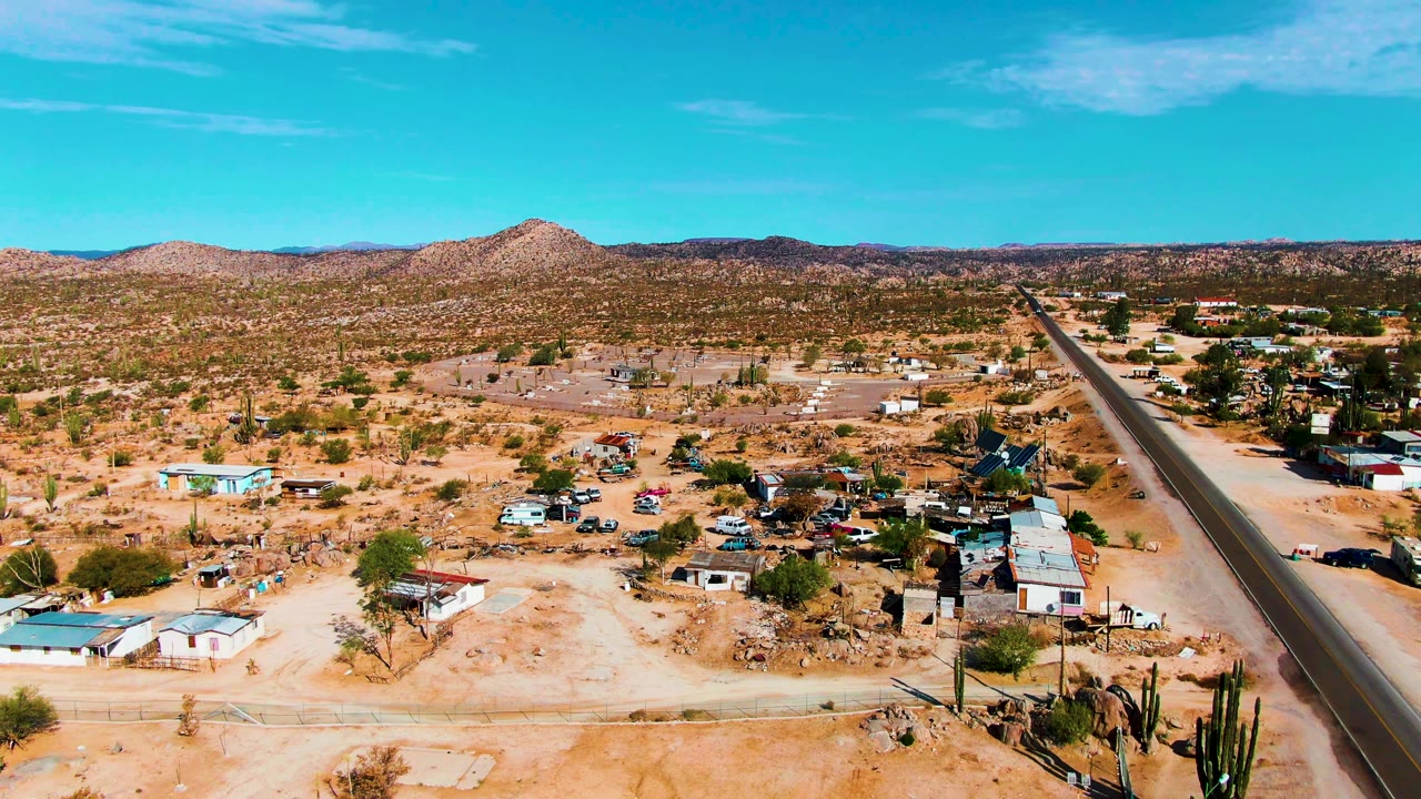 Aerial frame over Catavina, Mexico showing a wider spread of sandy desert lots, small roadside structures, tall cactus silhouettes near the bottom, a dark paved road, and no visible people.