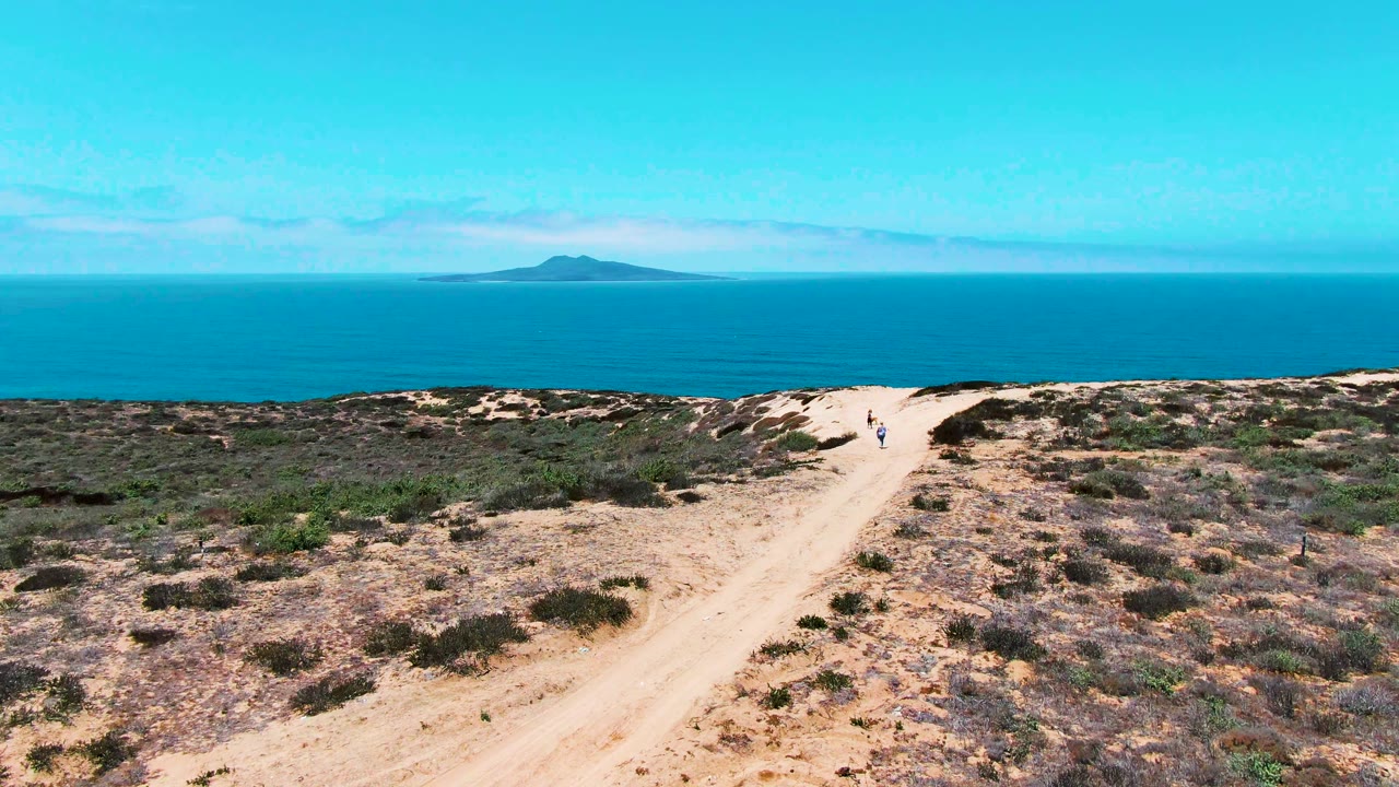 Wide elevated view in San Quintin BC Mexico showing two people on a sandy dirt path, dry scrub in the foreground, blue ocean beyond, and a small island low on the horizon.