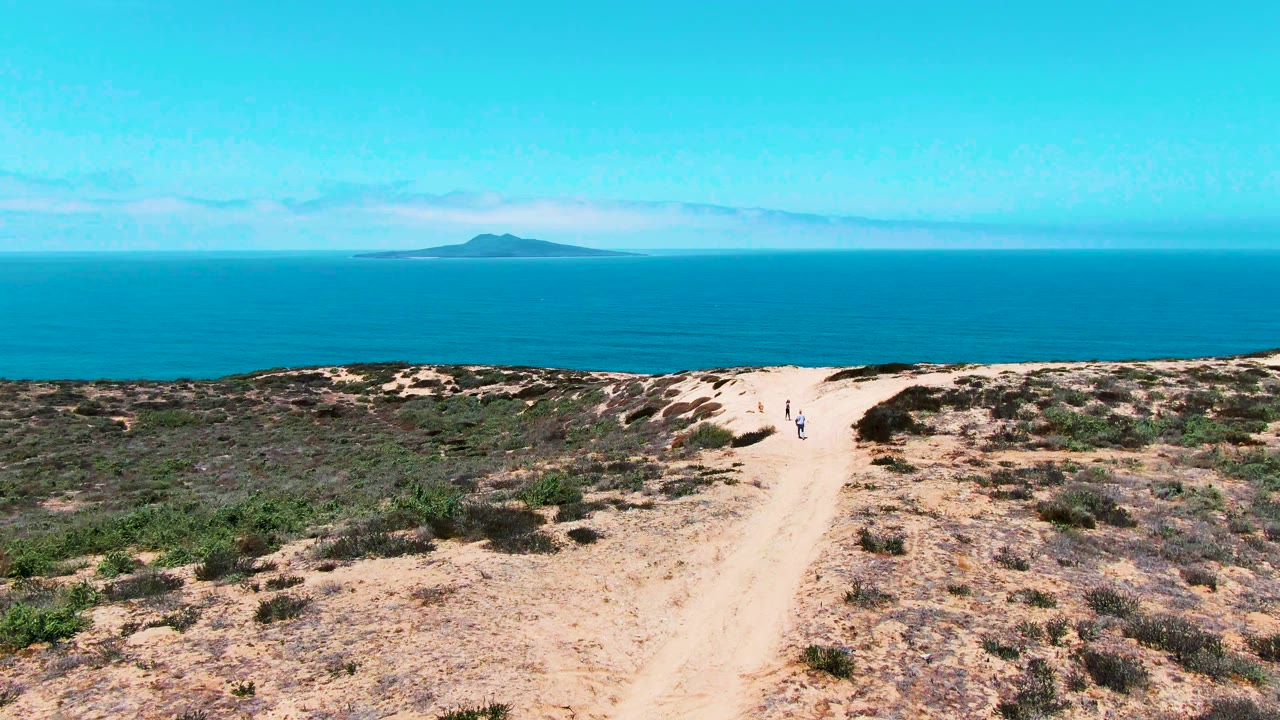 Wide view of San Quintin BC Mexico with two small walkers on the sandy path, scrubby terrain across the lower frame, calm blue water behind it, and a distant island near the left horizon.
