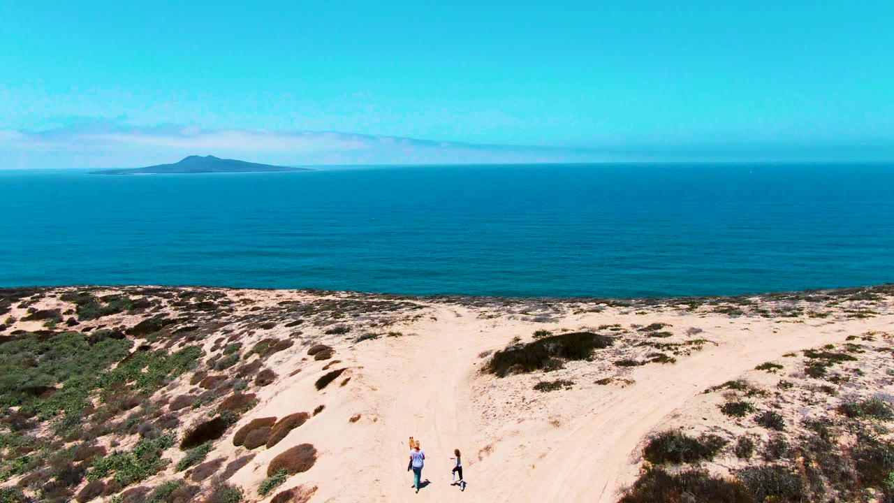 Two people walk on a pale dune path in San Quintin BC Mexico, with scrub-covered sand, blue ocean, a flat horizon, and a distant island behind them.